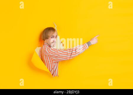 Pointage, affichage. Un jeune homme caucasien gai pose dans un fond de papier jaune déchiré, émotif et expressif. Se briser, se casser. Concept d'émotions humaines, expression faciale, ventes, publicité. Banque D'Images