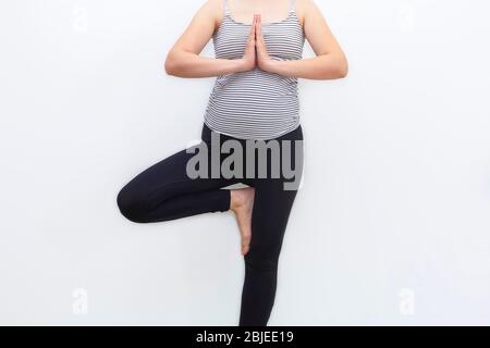 Une femme enceinte non reconnaissable qui fait du yoga prénatal dans l'arbre pose sur fond blanc neutre Banque D'Images