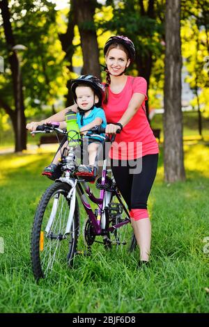 Un petit enfant dans un casque de protection sourit assis sur le siège de vélo d'un enfant sur un grand vélo de montagne avec sa mère sur le fond de l'herbe verte Banque D'Images