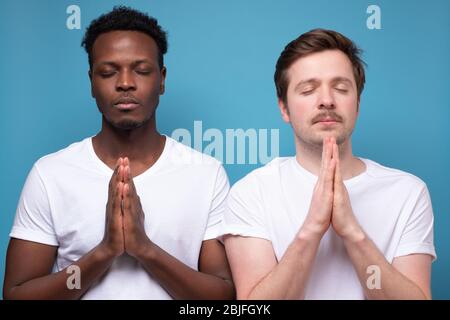 Des hommes adultes pleins d'espoir qui ferment les yeux et qui pressent les paumes ensemble prient, espérant le meilleur. Calme calme les gars tenant les mains dans namaste tout en méditant Banque D'Images