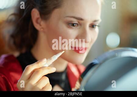 Gros plan sur la femme dans le salon en journée ensoleillée regardant dans le miroir et en utilisant le correcteur oculaire liquide. Banque D'Images