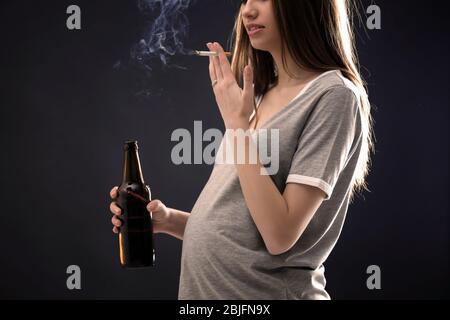 Femme enceinte fume la cigarette et boit la bière sur fond sombre Banque D'Images