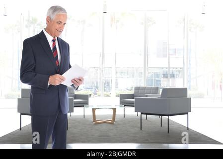 Un cadre d'affaires mûr debout dans un hall de bureau moderne lecture des documents qu'il tient dans ses mains. Banque D'Images