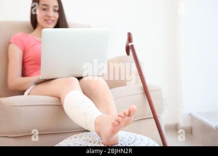 Belle jeune femme avec ordinateur portable et jambe en bandage assis sur un canapé à la maison Banque D'Images