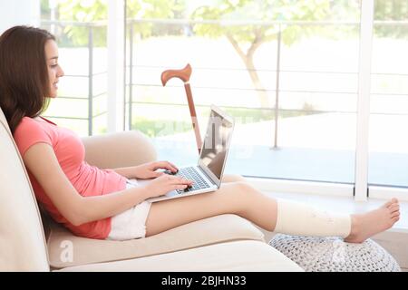 Belle jeune femme avec ordinateur portable et jambe en bandage assis sur un canapé à la maison Banque D'Images