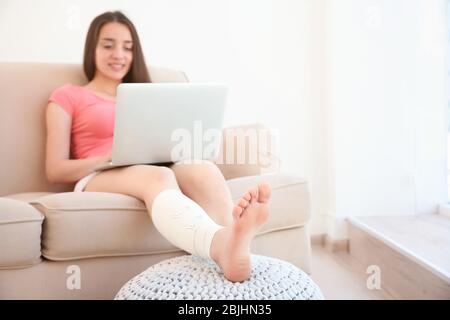 Belle jeune femme avec ordinateur portable et jambe en bandage assis sur un canapé à la maison Banque D'Images