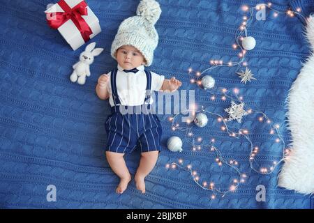 Joli petit bébé dans un chapeau avec jouet, cadeau, lumières de Noël et décorations sur tissu tricoté Banque D'Images