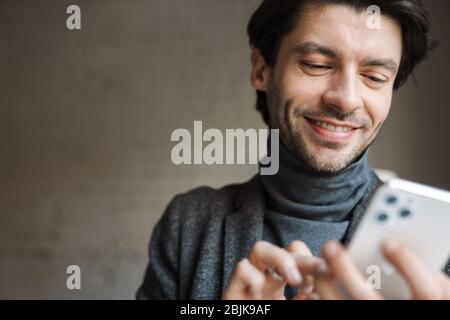 Gros plan sur un jeune homme souriant portant un turtleneck à l'intérieur, à l'aide d'un téléphone portable Banque D'Images