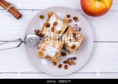 Tranches de strudel à tarte aux pommes maison dans une assiette avec des ingrédients sur une table rustique en bois blanc. Tarte aux pommes, aux raisins secs et à la cannelle, pâtisseries sucrées Banque D'Images