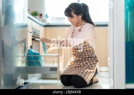 Jolie jeune femme asiatique qui charge le lave-vaisselle dans des placards de cuisine tout en faisant le nettoyage à la maison pendant le séjour à la maison en utilisant le temps libre abou Banque D'Images