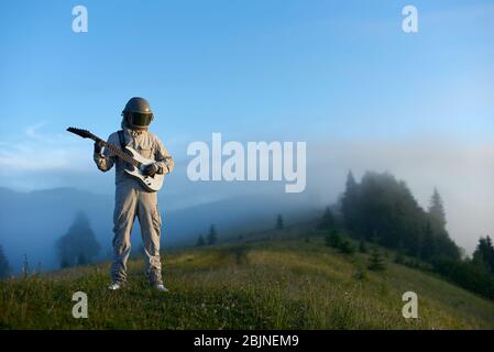 Voyageur de l'espace portant un costume et un casque, jouant de la guitare, debout sur la glade montagne verte ensoleillée le matin, les collines brumeuses et le ciel bleu sur fond. Concept d'astronautique, de musique et de nature. Banque D'Images