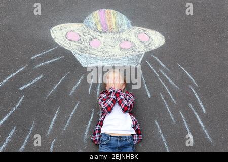 Petite fille allongé près de craie dessin de vaisseau spatial étranger sur l'asphalte Banque D'Images