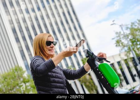 Belle femme élégante scannant le code qr avec son téléphone pour louer un scooter électrique avec gratte-ciel bâtiment en arrière-plan Banque D'Images