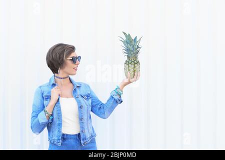 Jolie fille hipster posant avec de l'ananas près du mur blanc Banque D'Images