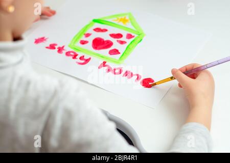 L'enfant écrit des mots rester maison avec l'aquarelle rouge sous la maison dessinée avec des coeurs sur feuille de papier. Concept de séjour à la maison. Banque D'Images