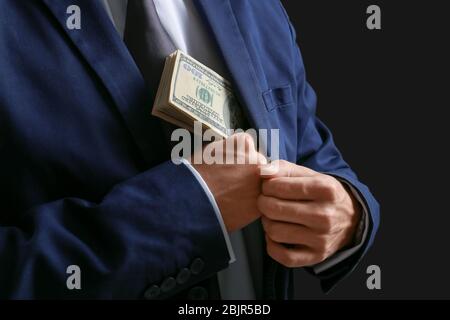 Homme en costume formel mettant de l'argent en poche sur fond sombre, closeup Banque D'Images