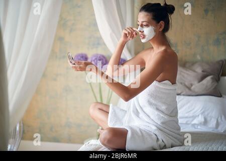 Jeune fille gaie assise dans le lit et regardant le miroir.Beauté et le concept de soins du corps. Banque D'Images