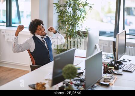 le jeune homme d'affaires afro-américain s'étire avec concensment sur son lieu de travail dans son bureau après avoir reçu une bonne nouvelle Banque D'Images