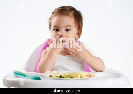 Bébé manger avec les mains sur la chaise haute isolée dans le fond blanc Banque D'Images