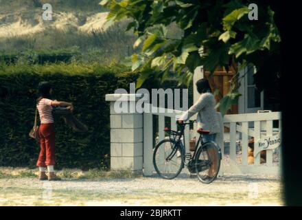 Deux jeunes femmes, l'une tenant un panier l'autre avec son style néerlandais s'asseoir à vélo, en parlant à l'extérieur d'une maison à St Cast le Guildo, Bretagne, France 1974. Banque D'Images