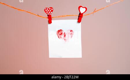 un morceau de papier avec un coeur à motifs de traces de doigts de clothespins rouges avec un coeur sur une corde. Amour de la Saint-Valentin. Fond rose. Espace de copie. Banque D'Images