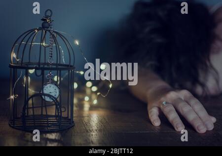 Photo conceptuelle de l'emprisonnement pendant l'accouchement du coronavirus. Une horloge emprisonnée dans une cage d'oiseaux avec une femme dépressive caucasienne posée sur une table Banque D'Images