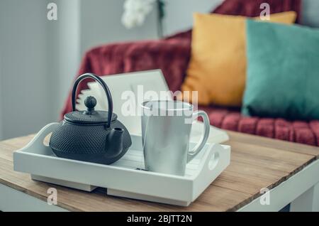 Gros plan et mise au point sélective sur un plateau avec une théière et une tasse sur une table en bois. Couverture bordeaux, coussins jaunes et verts sur un canapé gris dans le bl Banque D'Images