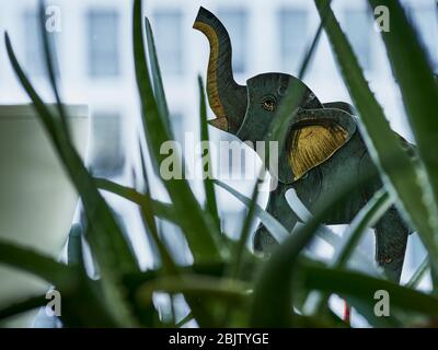 Jouet éléphant peint avec des plantes en premier plan et des fenêtres de ville en arrière-plan. Concept: Safari intérieur Banque D'Images