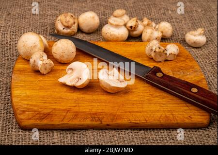 Moitiés de jeunes champignons coupés et un couteau avec poignée en bois sur un panneau de découpe en bois et champignons dispersés sur un fond de gros homespun fa Banque D'Images