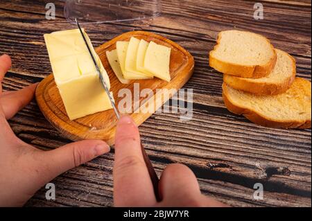 Quelqu'un coupe un peu de beurre avec un couteau d'un morceau de beurre dans un plat de beurre en bois, des tranches de fromage et du pain grillé de blé frais sur un fond en bois. C Banque D'Images