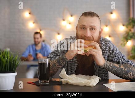 Homme barbu mangeant du hamburger délicieux dans le café Banque D'Images