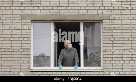 L'homme âgé reste dans la fenêtre dans le masque de protection. Isolé à domicile en quarantaine. Éclosion de virus. Grand-père aîné. COVID-19 prévention de la pandémie de coronavirus. Distanciation sociale. Concept de séjour à la maison Banque D'Images