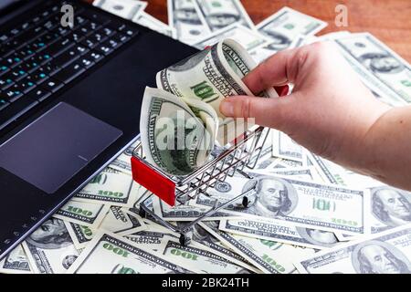 La main féminine met des dollars dans un panier. Ordinateur portable et dollars sur la table. Banque D'Images