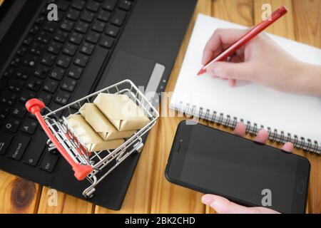 Concept de maquette d'achat en ligne. Fille prend des notes à un ordinateur portable avec téléphone portable en main et un petit panier avec des boîtes sur l'ordinateur. Banque D'Images