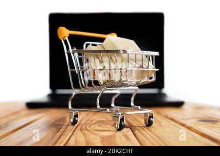 Mini supermarché panier plein de boîtes d'expédition se tenir sur une table en bois contre un ordinateur portable noir flou sur fond blanc. Banque D'Images