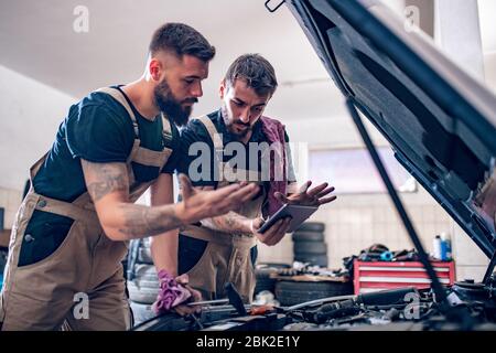 Deux mécaniciens travaillant sur des rapports dans un atelier.Mécanique avec outil d'acquisition diagnostiquant la voiture dans un capot ouvert. Banque D'Images