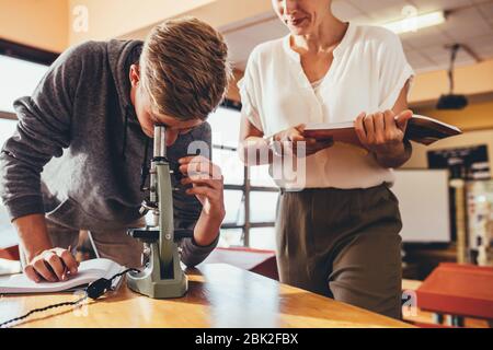 Étudiant en lycée travaillant dans une classe scientifique avec son professeur debout par. Garçon regardant les lames en classe de biologie à travers un microscope. Banque D'Images