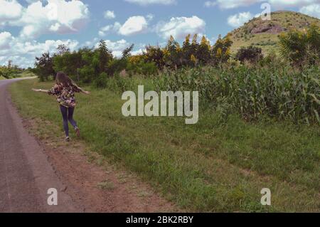 Route africaine étonnante, paysages verts avec montagnes, champs de maïs, fleurs jaunes fleuries. La femme qui courant depuis l'arrière suit la route vers Banque D'Images