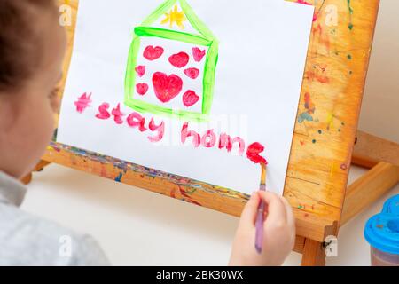 L'enfant écrit des mots rester maison avec l'aquarelle rouge sous la maison peinte avec des coeurs sur le chevalet. Concept de séjour à la maison. Banque D'Images