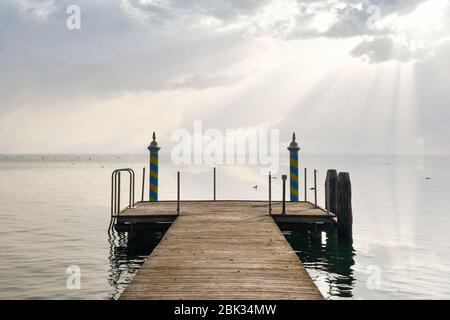 Une jetée en bois sur le lac avec un ciel spectaculaire et des rayons du soleil qui se diffusent à travers les nuages et se réfléchissent sur l'eau, lac de Garde, Vérone, Italie Banque D'Images