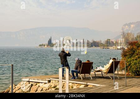 Un groupe de personnes assis sur des bancs et prenant des photos avec un smartphone sur le bord du lac, dans une journée d'hiver ensoleillée, Bardolino, Vérone, Vénétie, Italie Banque D'Images