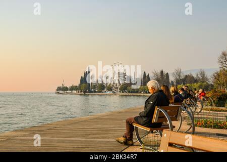 Homme âgé et autres personnes assises sur des bancs sur le lac avec une roue panoramique en arrière-plan au coucher du soleil, Bardolino, VeronaVeneto, Italie Banque D'Images