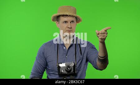 Portrait de l'homme de la mode photographe touristique pointant vers quelque chose avec son doigt. Beau homme en vacances en chemise bleue et chapeau avec appareil photo rétro. Placez pour votre logo. Clé Chroma. Écran vert Banque D'Images