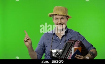 Portrait de l'homme de mode photographe touristique avec valise en main pointant quelque chose avec son doigt. Beau homme en vacances en chemise bleue et chapeau avec appareil photo rétro. Clé Chroma Banque D'Images