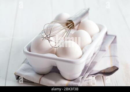 Œufs de poulet de ferme bio fraîchement cueillis dans un plateau en céramique sur fond de bois avec fouet en métal et serviette en tissu, espace de copie. Une alimentation naturelle saine Banque D'Images