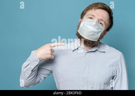 L'homme pointe l'index vers le masque médical sur son visage, ce qui n'est pas ajusté en raison de la barbe taillée. Concept de quarantaine. Banque D'Images