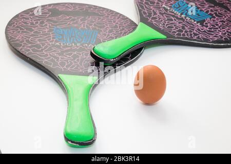 Un œuf repose et remplace la balle au milieu entre deux raquettes de tennis de plage sur fond blanc, la composition exprime le concept de Banque D'Images