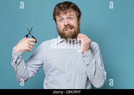 Shaggy homme avec une barbe et des ciseaux dans les mains, alarmé qu'il devra couper ses cheveux indépendamment pendant la quarantaine. Banque D'Images