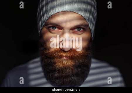 Portrait en gros plan d'un homme barbu d'âge moyen avec des yeux expressifs et des ombres rayées sur son visage avec un arrière-plan noir Banque D'Images