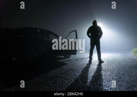 Un homme debout à côté d'une voiture, avec porte ouverte, garé sur le côté de la route, sous un feu de rue. Une nuit d'hiver brumeuse Banque D'Images